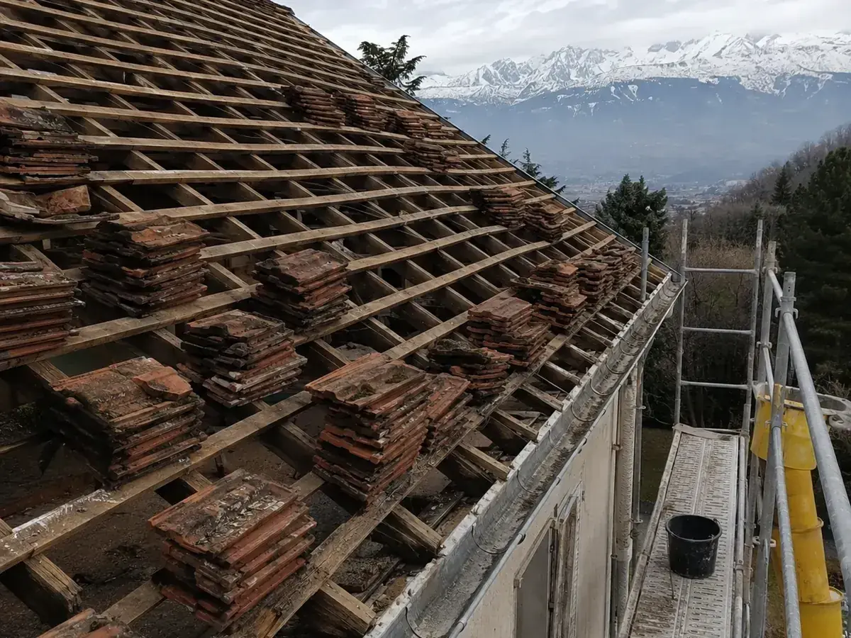 Versant de toiture mis à nu, anciennes tuiles empilées sur le liteaunage et l'échafaudage, massifs enneigés en arrière-plan