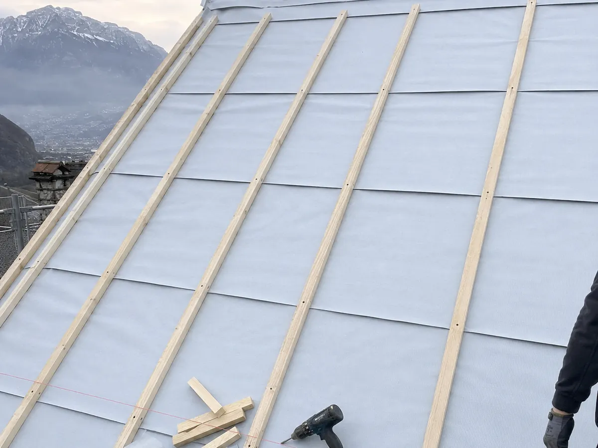 Écran sous-toiture gris posé sur un versant avec contre-liteaux en bois, visseuse posée dessus, montagnes en arrière-plan