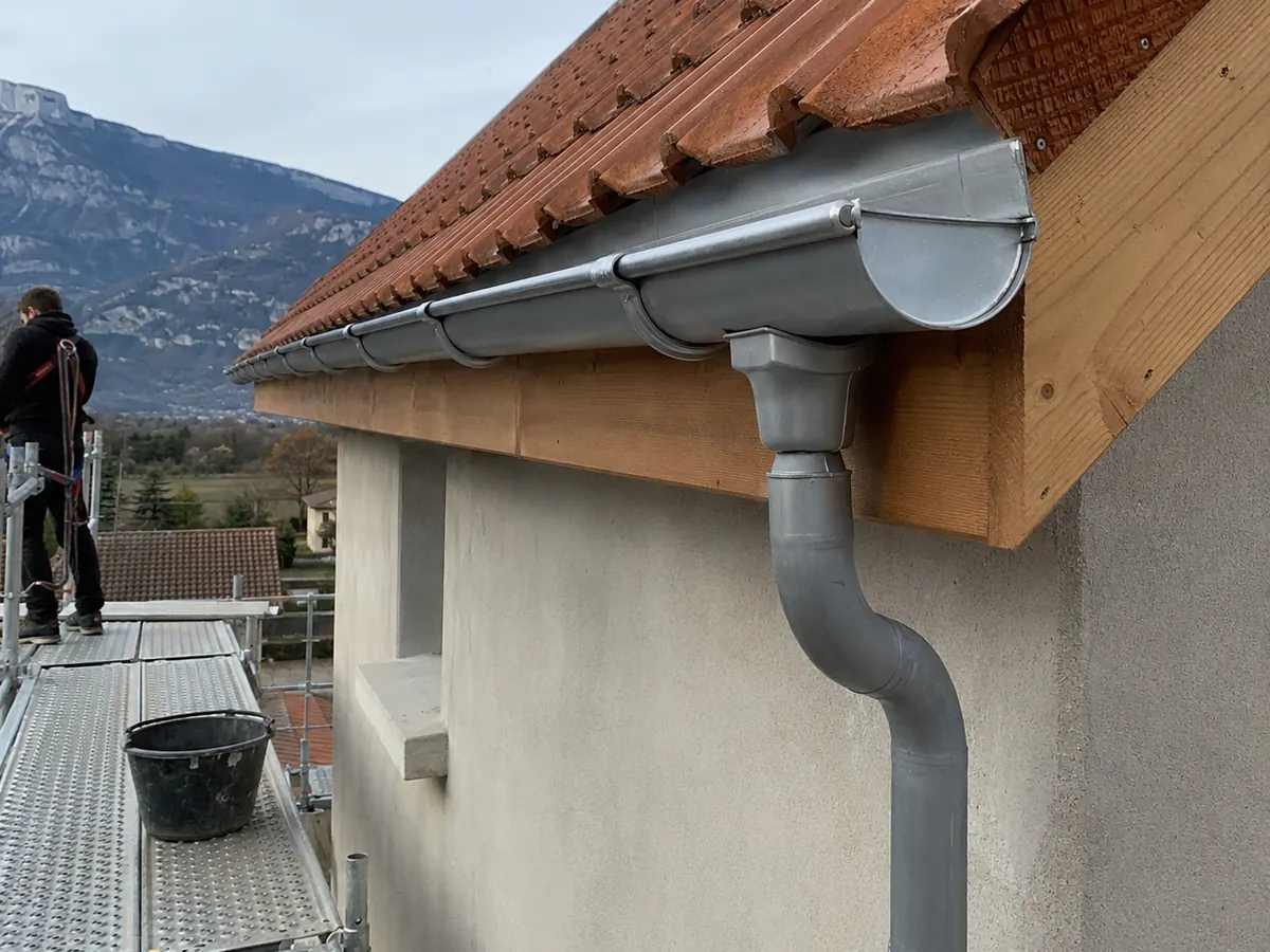 Gouttière et descente en zinc neuves sous un débord de toit en tuiles rouges, échafaudage et montagnes en arrière-plan